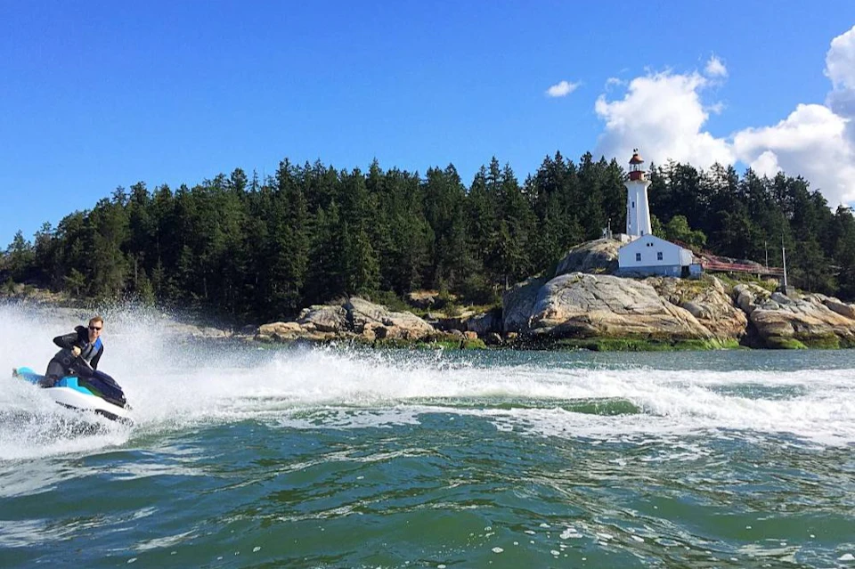 A white lighthouse on rocky shore surrounded by trees under a clear blue sky, with waves splashing in the foreground.