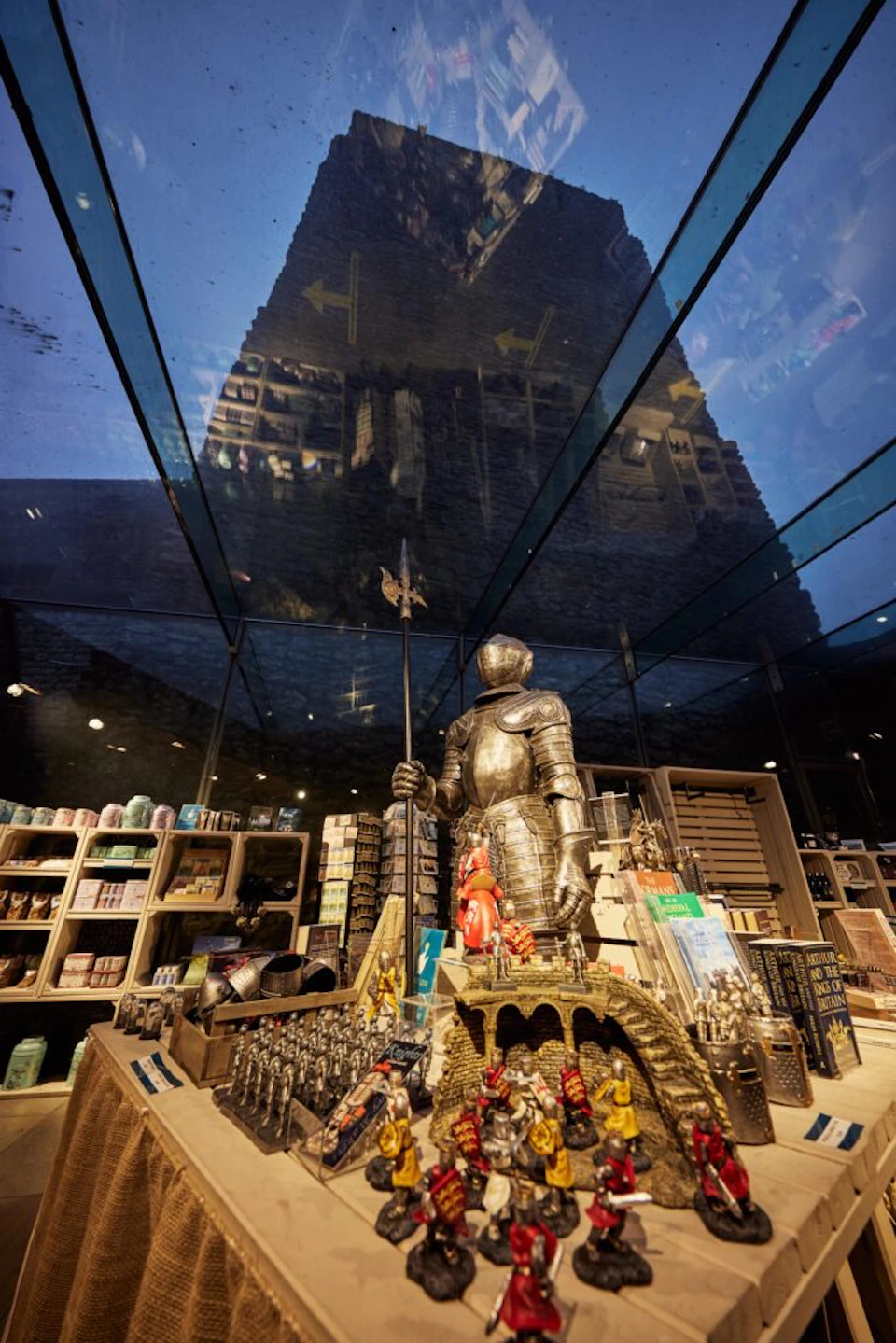 A store display features a knight statue surrounded by various medieval-themed merchandise, with a glass ceiling reflecting a castle.