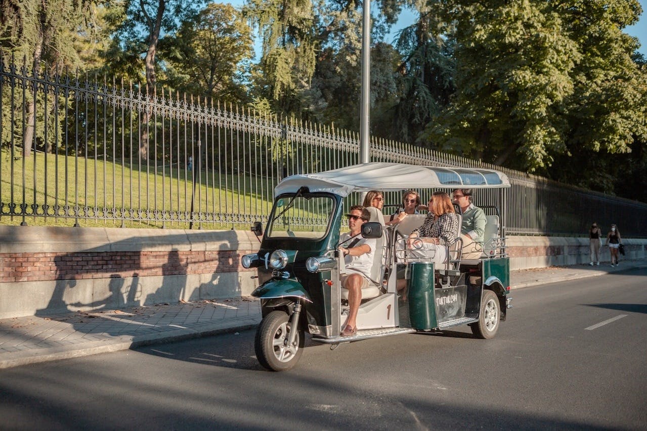 A group of people ride in a green tuk-tuk on a sunny day, traveling along a tree-lined street with a wrought iron fence on the side.
