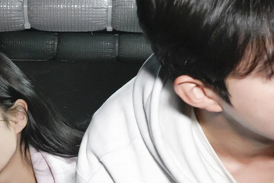 Two people wearing hoodies crawl through a dark, confined space with padded walls and pipes lining the ceiling.