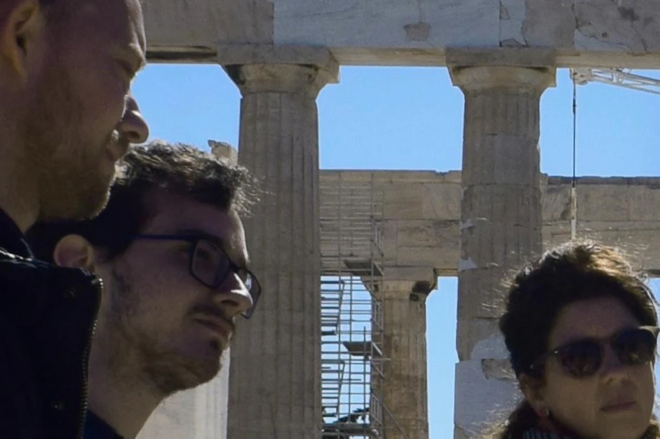 A group of people stands near the Parthenon ruins under a clear blue sky, engaging in a discussion or tour.