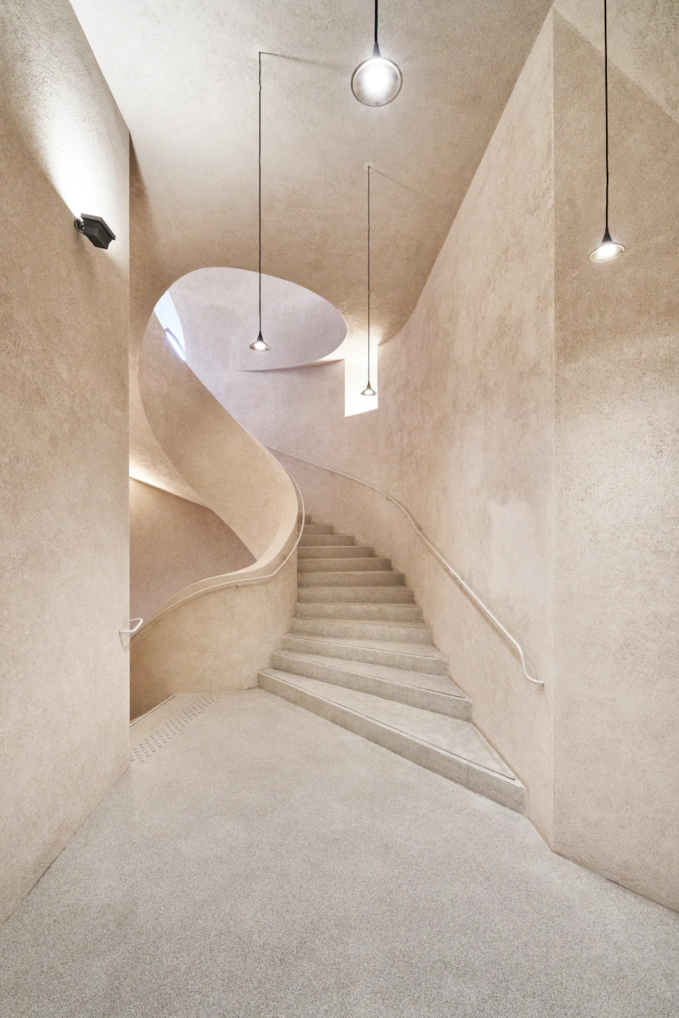 A minimalist, curving staircase in a beige-toned interior with three pendant lights hanging from the ceiling.