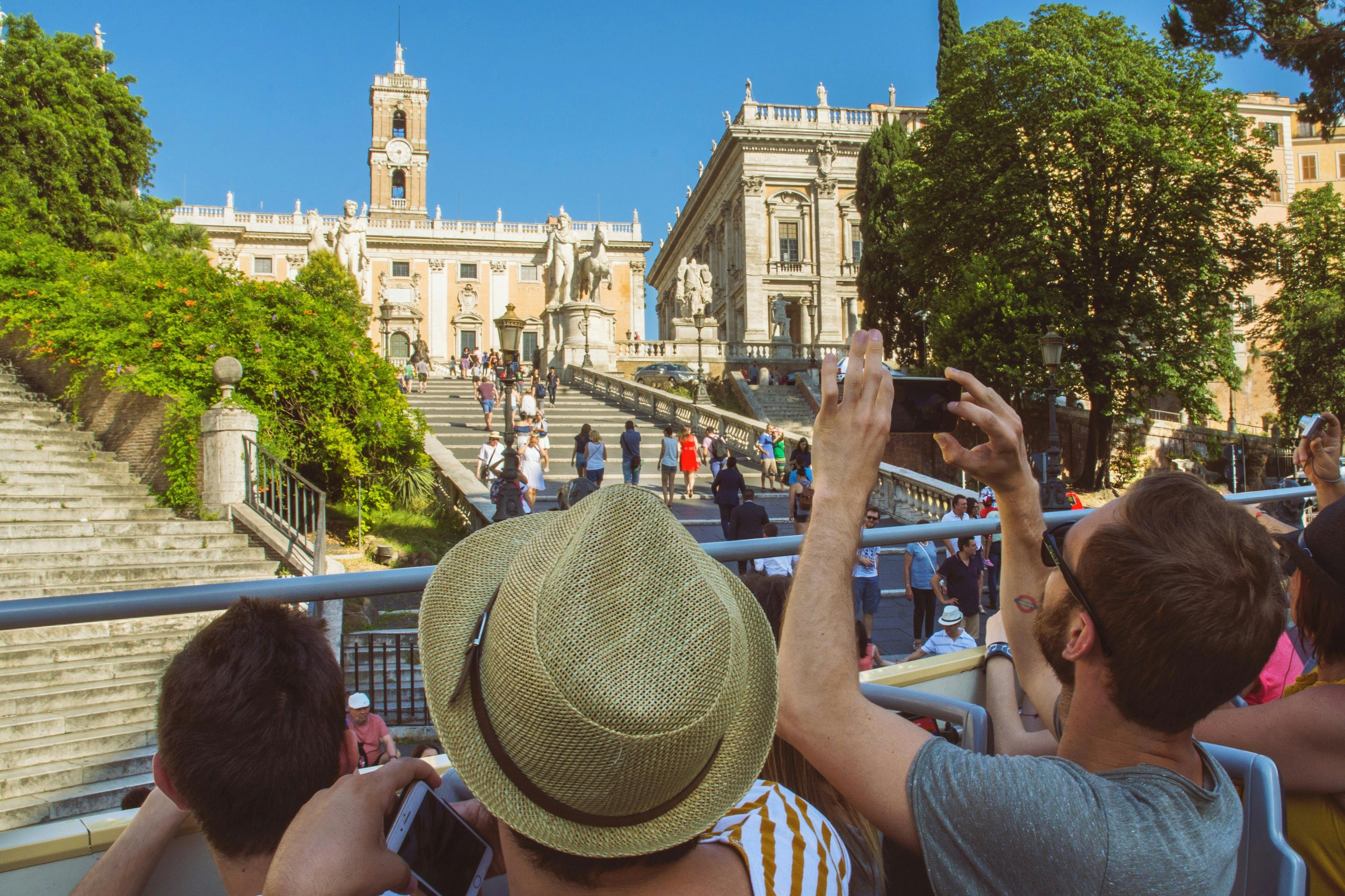 Tourists taking photos of a historical building with a grand staircase, statues, and lush greenery on a sunny day.