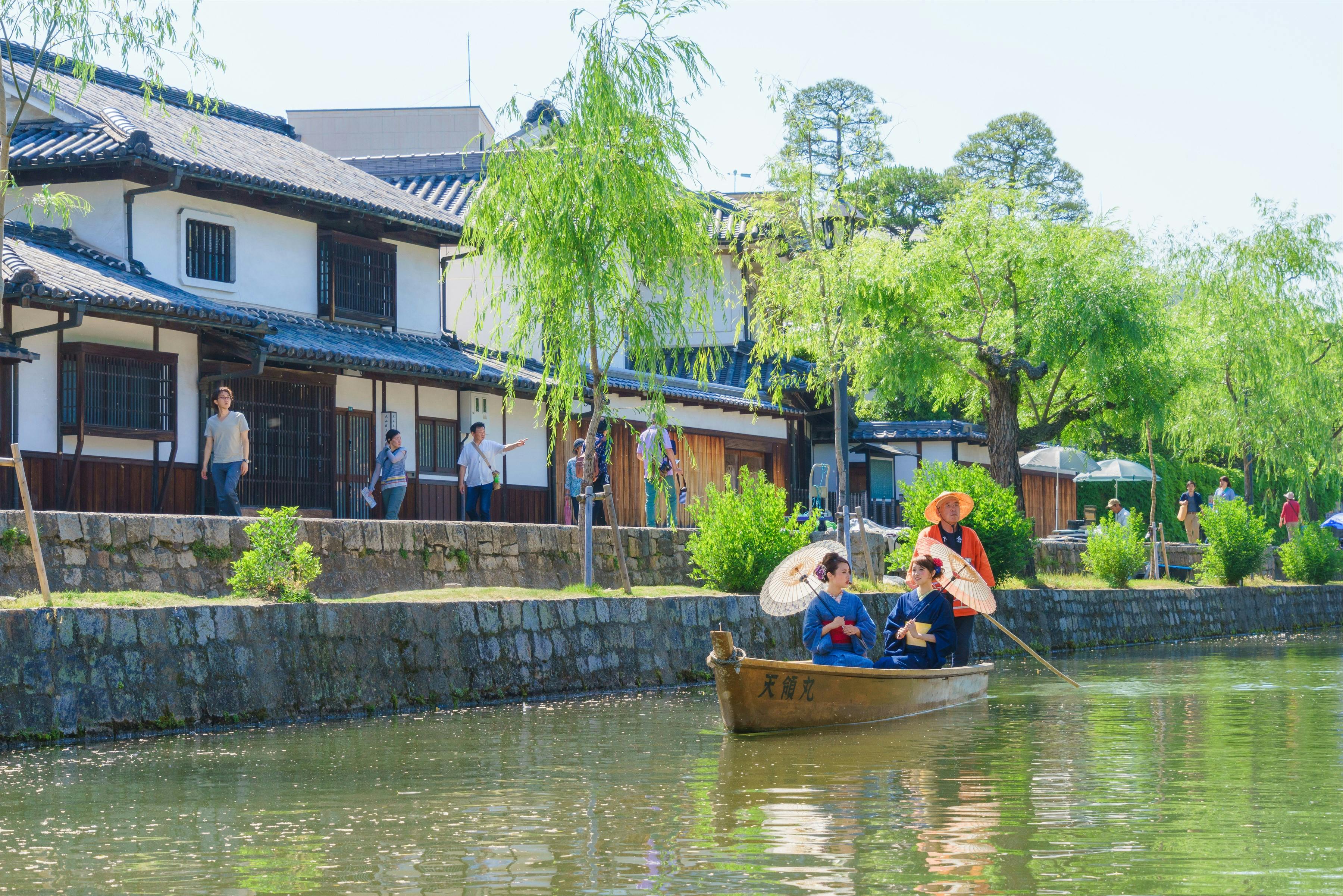 Un bateau en bois avec des personnes en vêtements traditionnels sur un canal, avec des spectateurs marchant près d'un bâtiment traditionnel et d'arbres en arrière-plan.