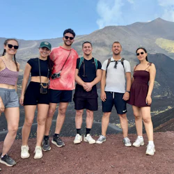 Six people in casual hiking attire stand together on a rocky terrain with a mountain landscape in the background.