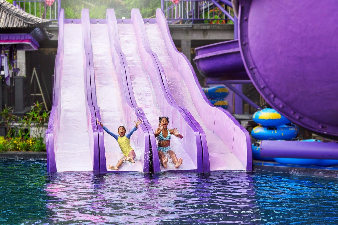 Two children, one in a yellow top and the other in a swimsuit, sliding down parallel purple water slides into a pool.