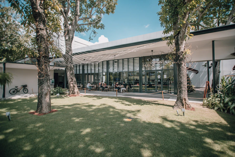 Outdoor seating area of a modern cafe with glass walls, surrounded by trees and grass, under a sunny sky.