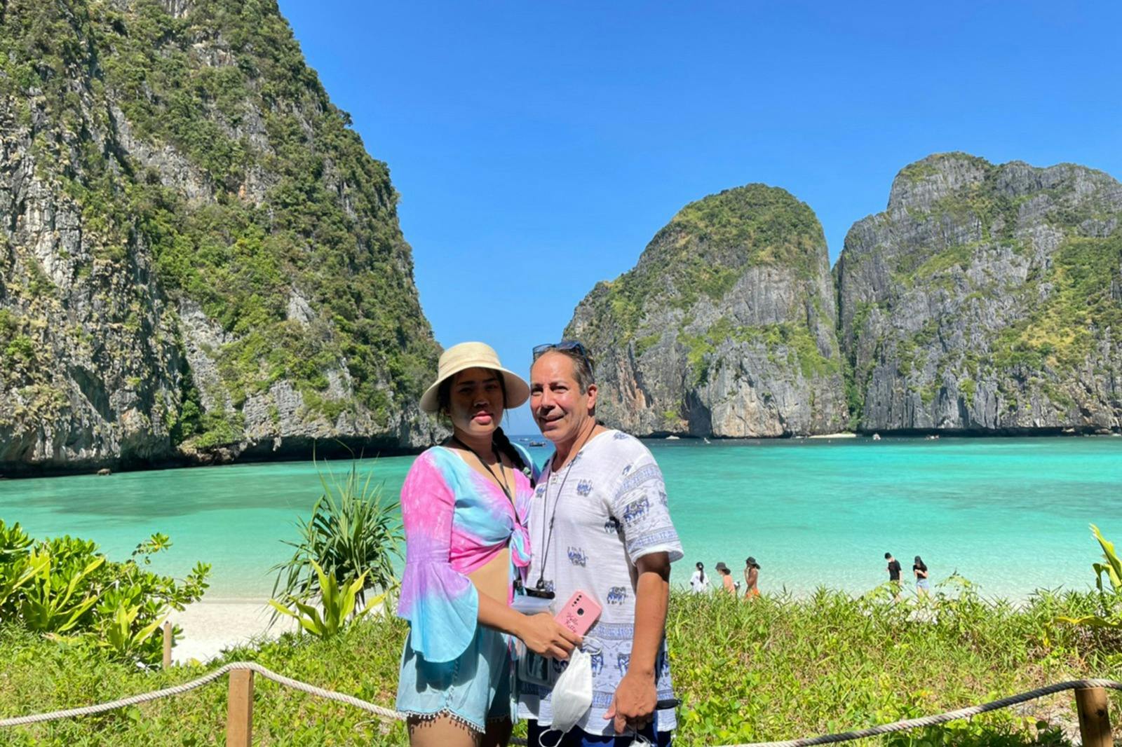 Pár stojí před tropickou pláží s průzračnou tyrkysovou vodou a velkými skalnatými útesy proti jasně modré obloze.