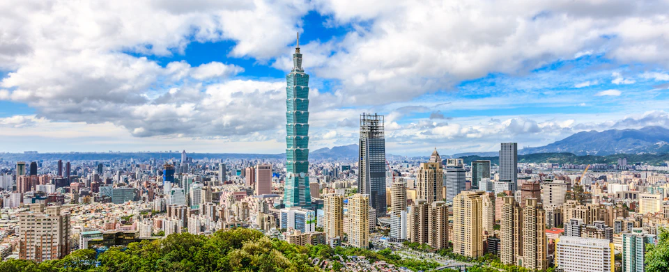 A skyline view of Taipei featuring the prominent Taipei 101 skyscraper among other buildings under a partly cloudy sky.
