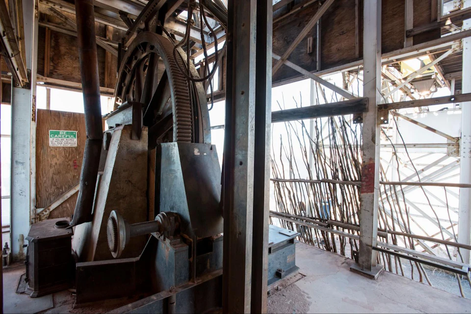 An industrial setting with large gears and machinery, surrounded by metal structures and a view of outdoor wooden barriers.