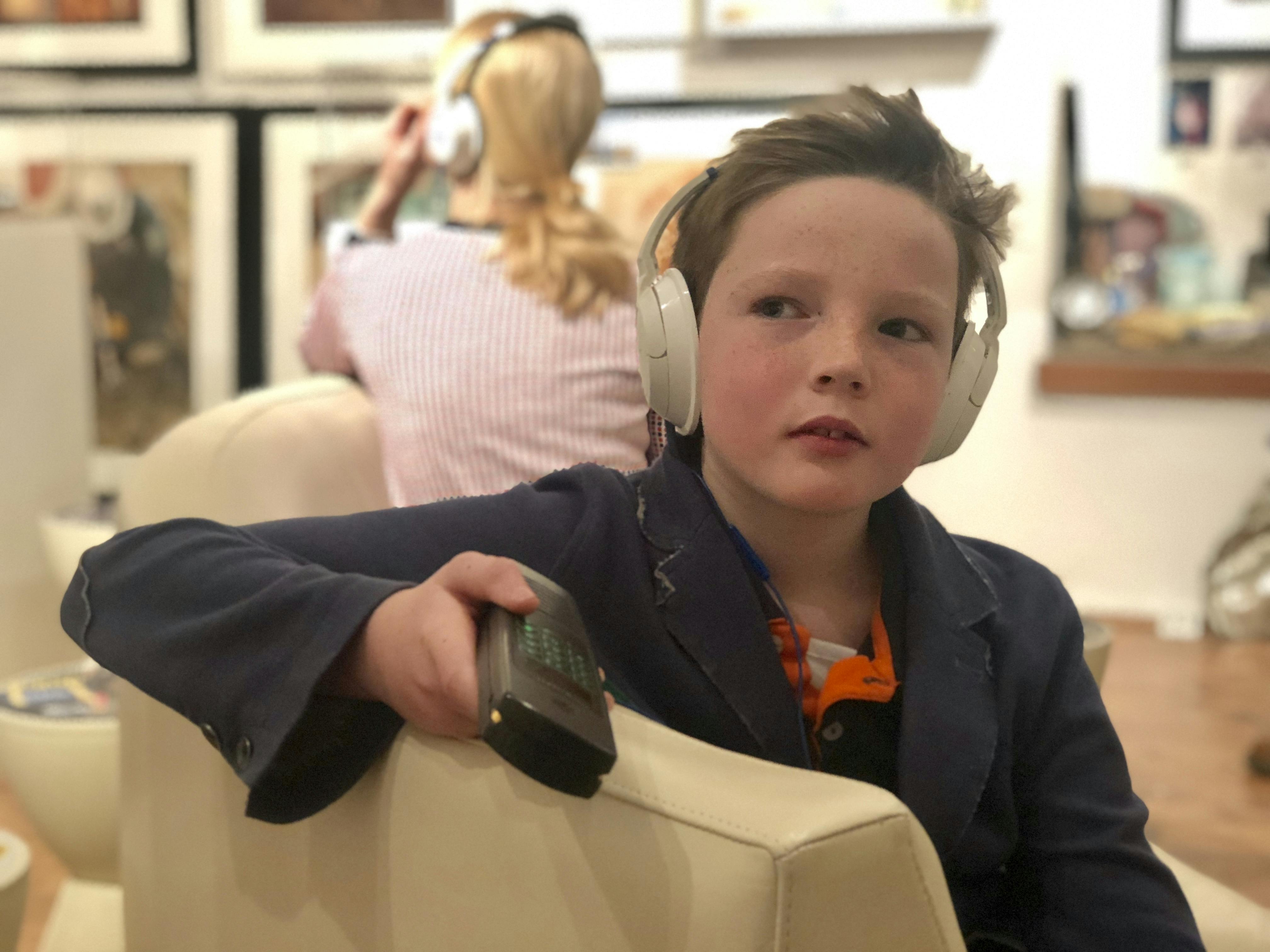 A young boy with headphones holds a remote while sitting indoors. An adult in the background also wears headphones.