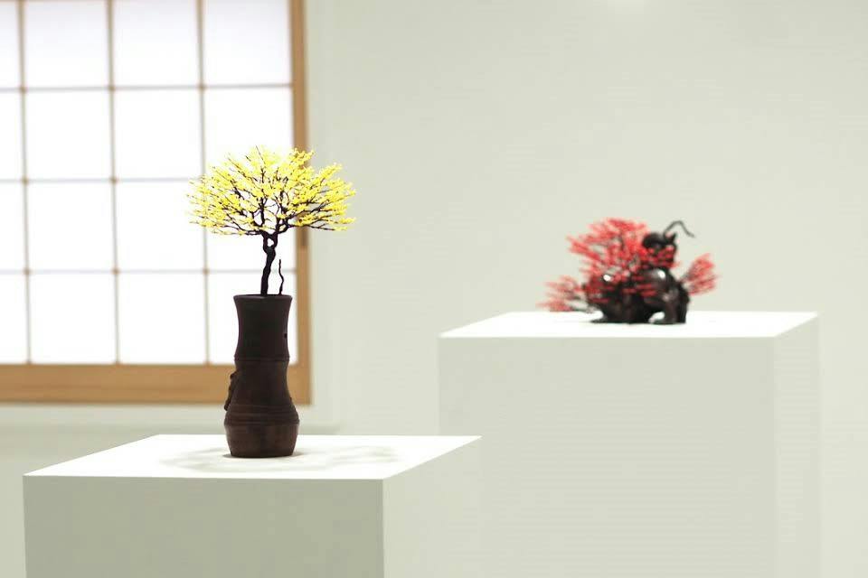 A yellow bonsai tree in a dark vase on a pedestal, with a red bonsai tree in the background. Minimalist indoor setting.