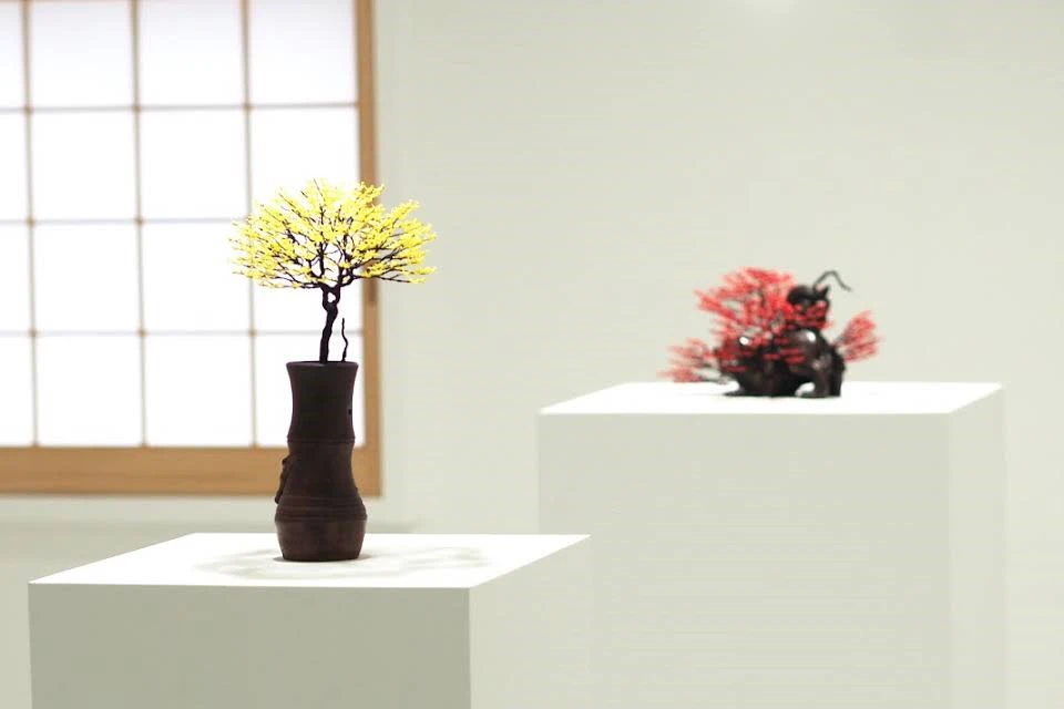 Two bonsai-style sculptures on display, one with yellow branches in a dark vase and another with red branches on a pedestal.