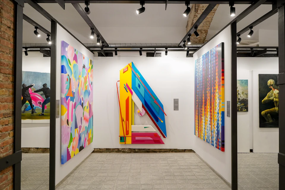 A gallery space featuring three colorful abstract paintings displayed on white walls with track lighting overhead.