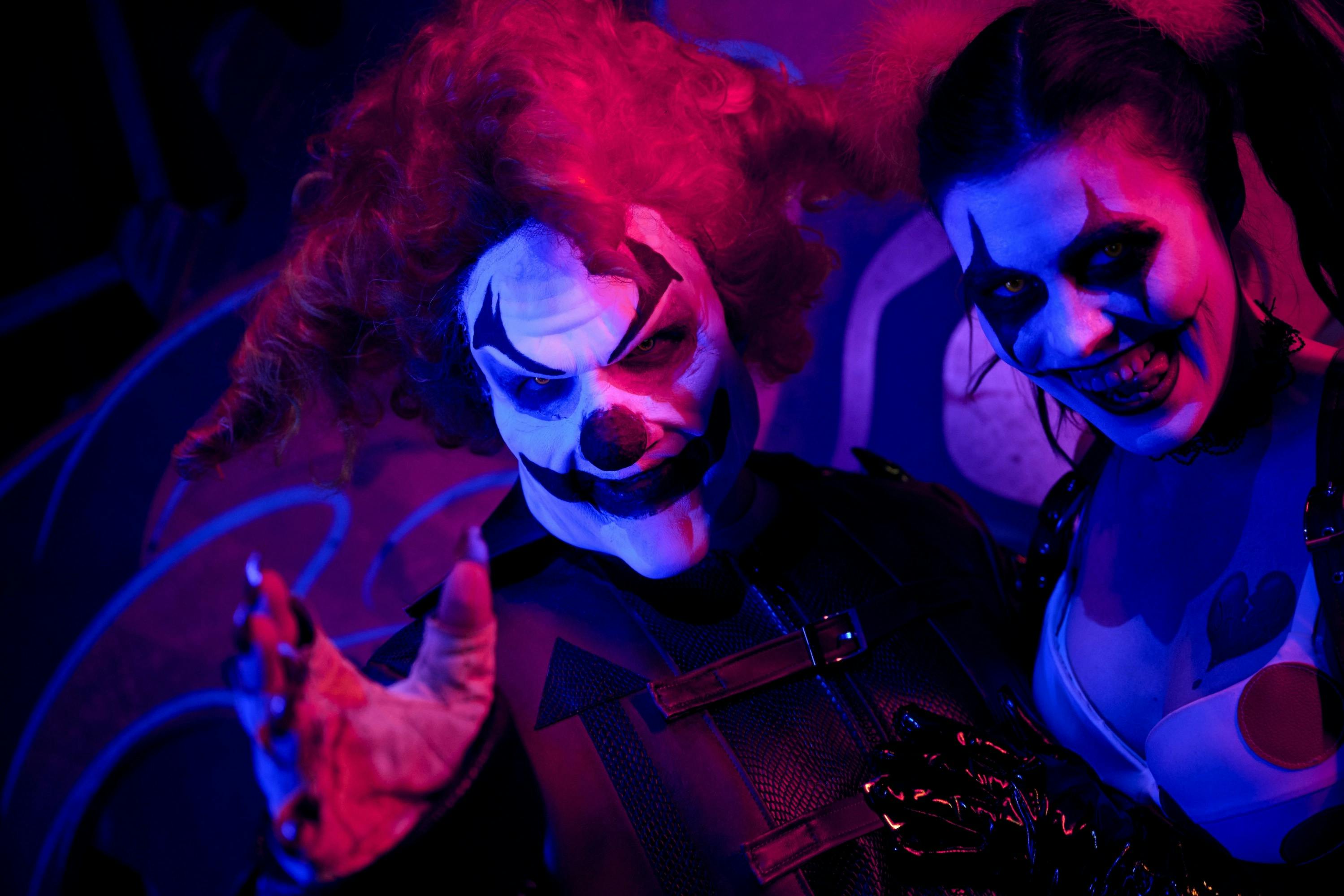 보라색과 파란색 조명으로 빛나는 소름끼치는 광대 메이크업과 의상을 입은 두 사람이 위협적으로 웃고 있습니다.