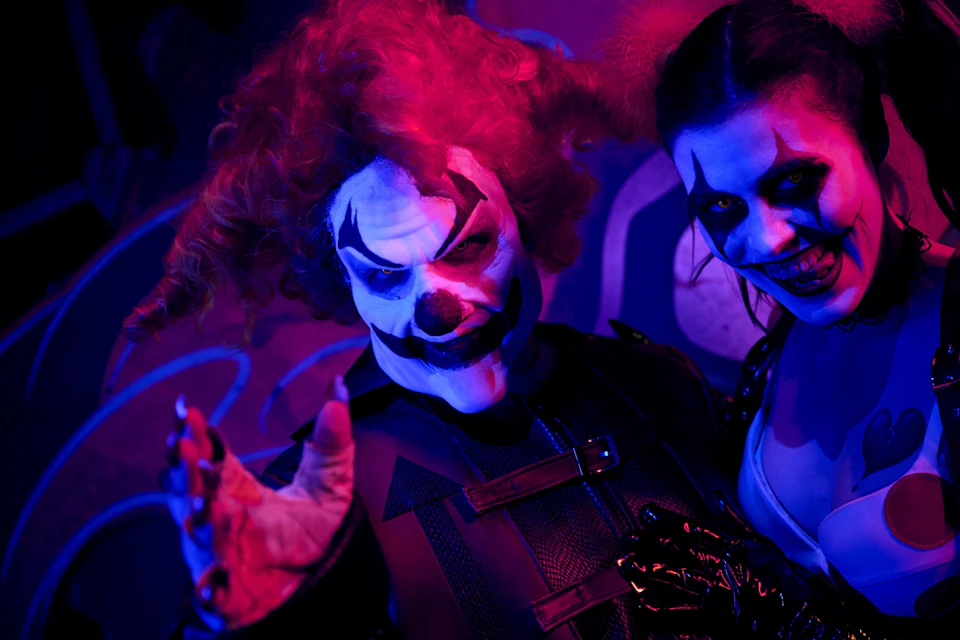 Two people in eerie clown makeup and costumes, illuminated by purple and blue lighting, smiling menacingly.