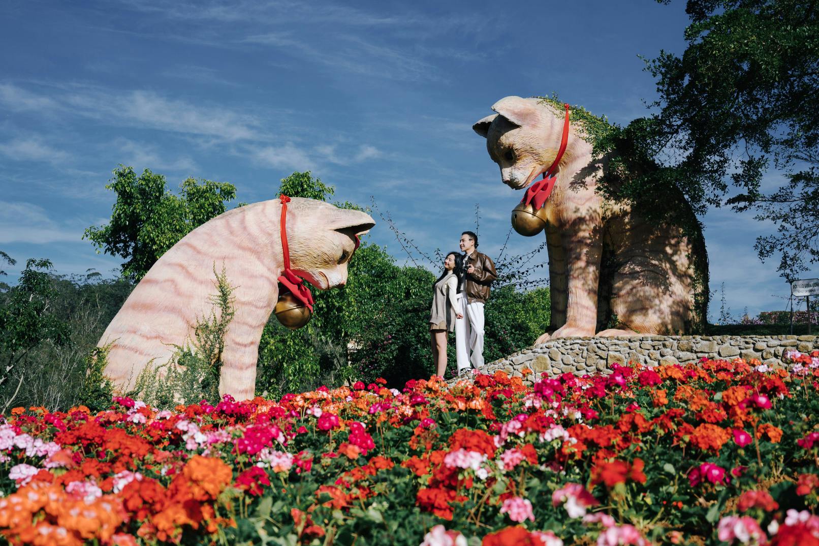 Two people standing among colorful flowers, with large cat statues wearing bells in a garden setting under a blue sky.