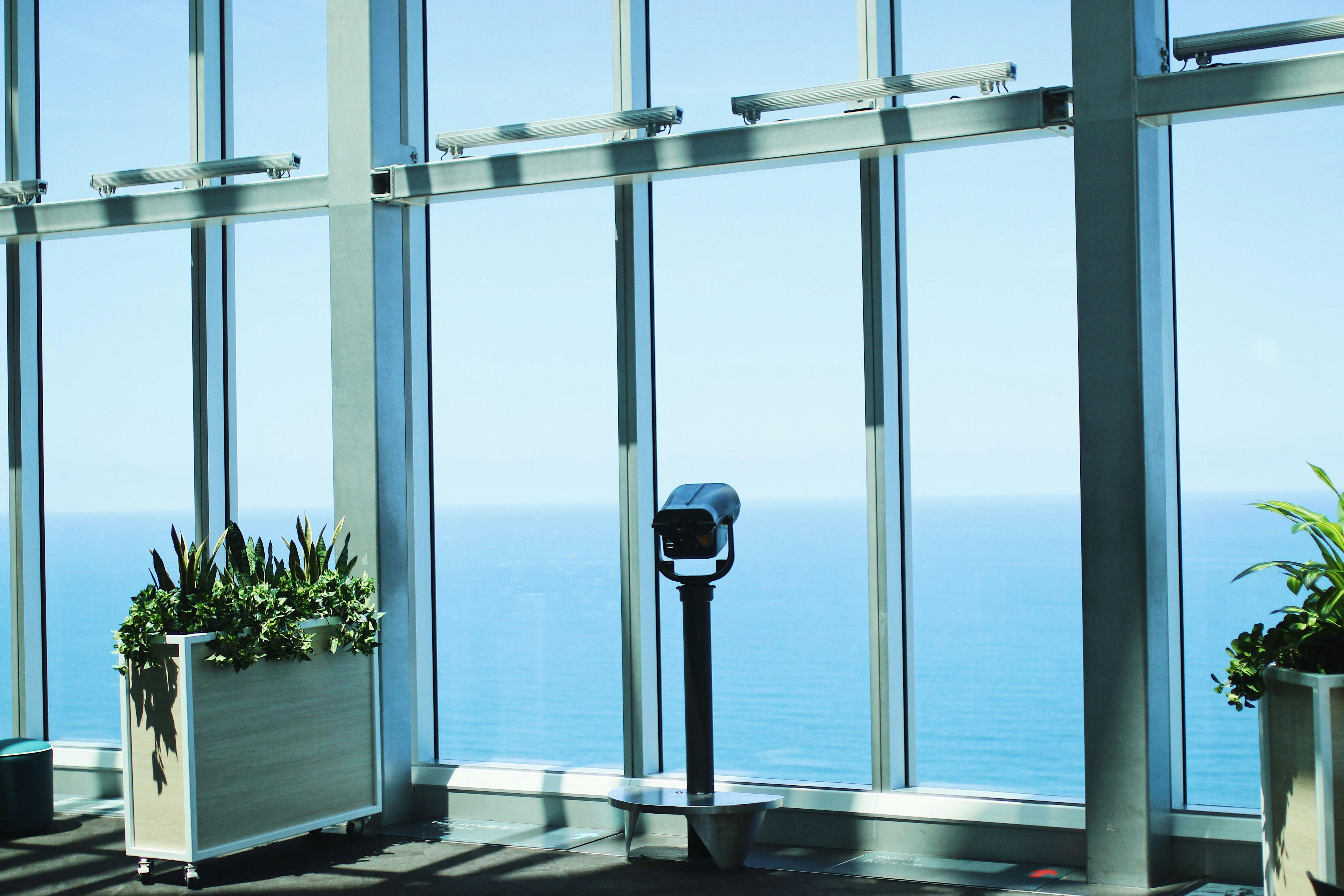Observation deck with a fixed binocular overlooking a clear blue ocean through large windows. Potted plants on the left.