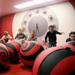 Barn i ett rum med röda och svarta spiralmönstrade bollar, en stor klocka på väggen och röd och beige inredning.