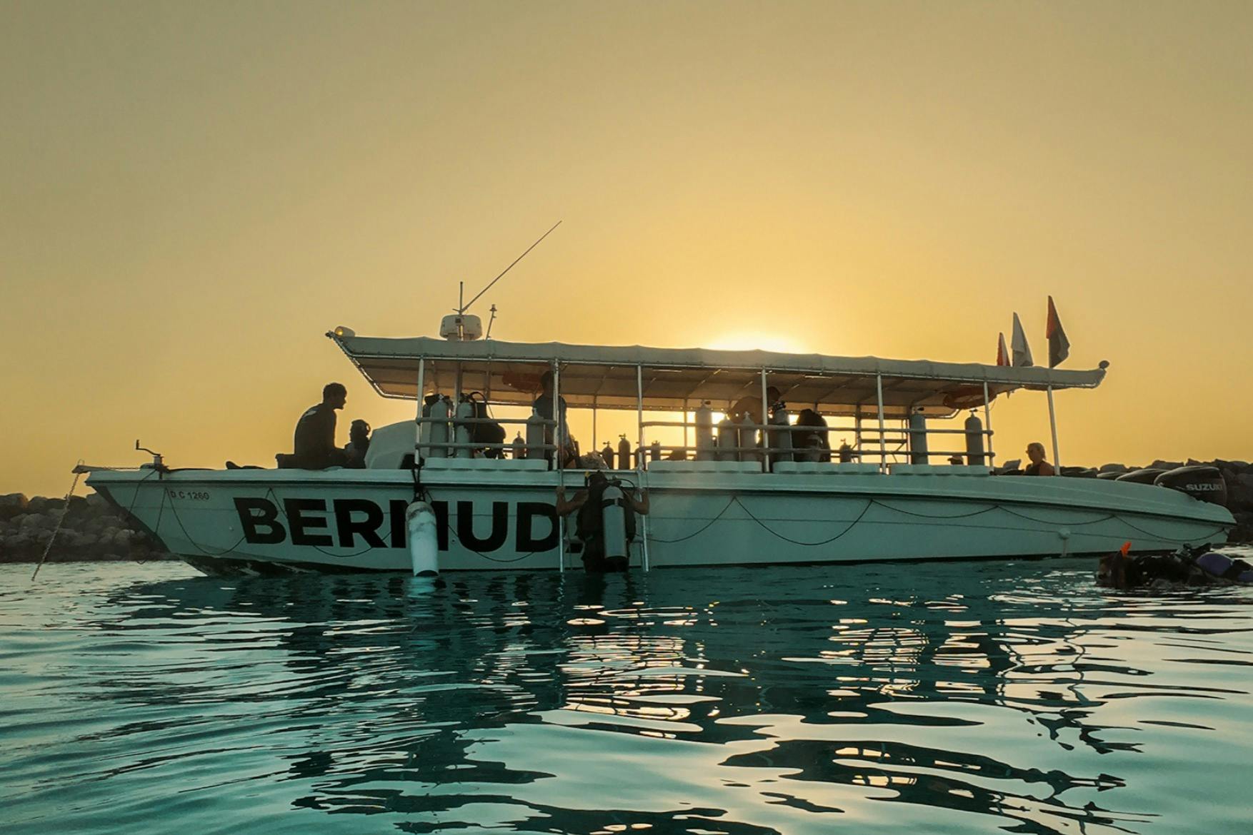 「バミューダ」と書かれたボートは、夕暮れ時の穏やかな水の上にあり、船上には人々のシルエットが見えます。