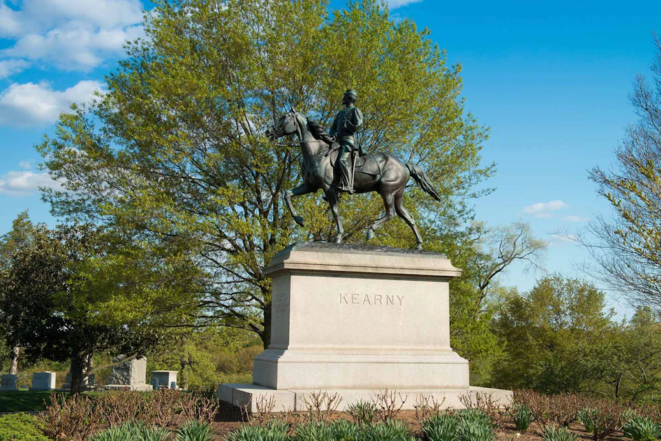 Equestrian statue of a rider named Kearny, set on a stone pedestal in a park with green trees and a blue sky.
