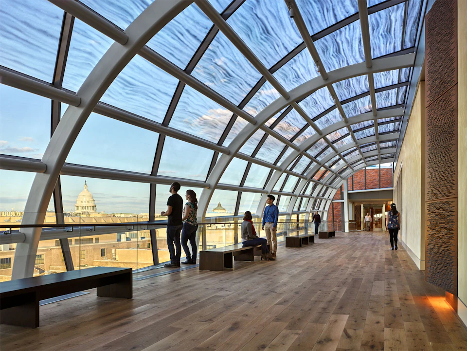 People stand and sit in a glass-enclosed hallway with a curved roof, offering views of a cityscape with a domed building in the distance.