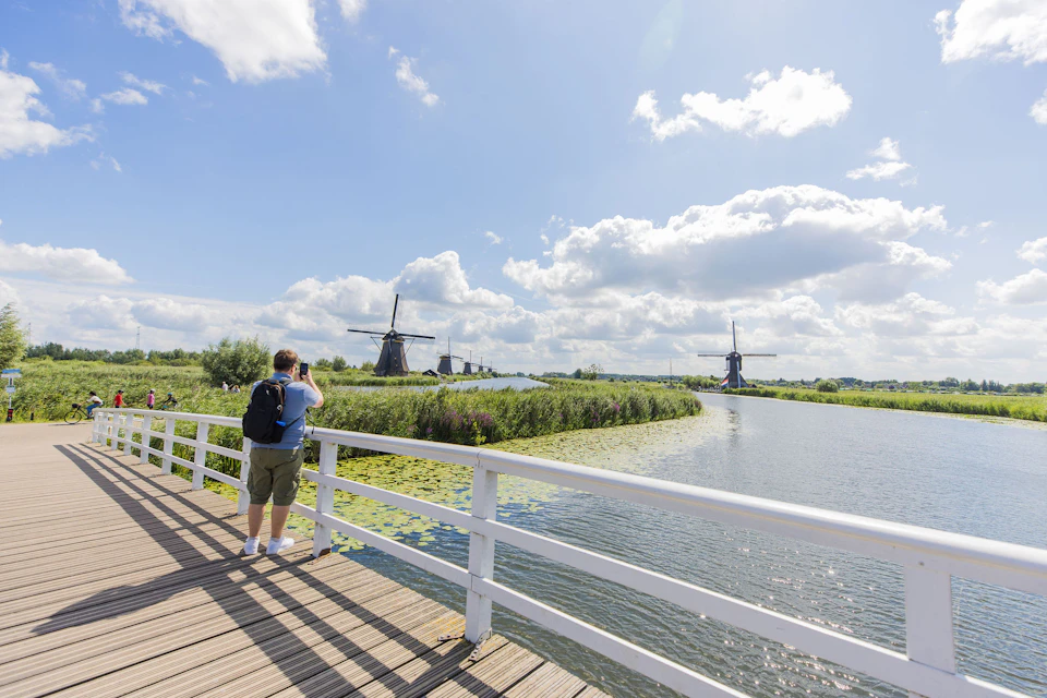 A person on a wooden bridge photographing a scenic view of windmills, water, and greenery under a partly cloudy sky.