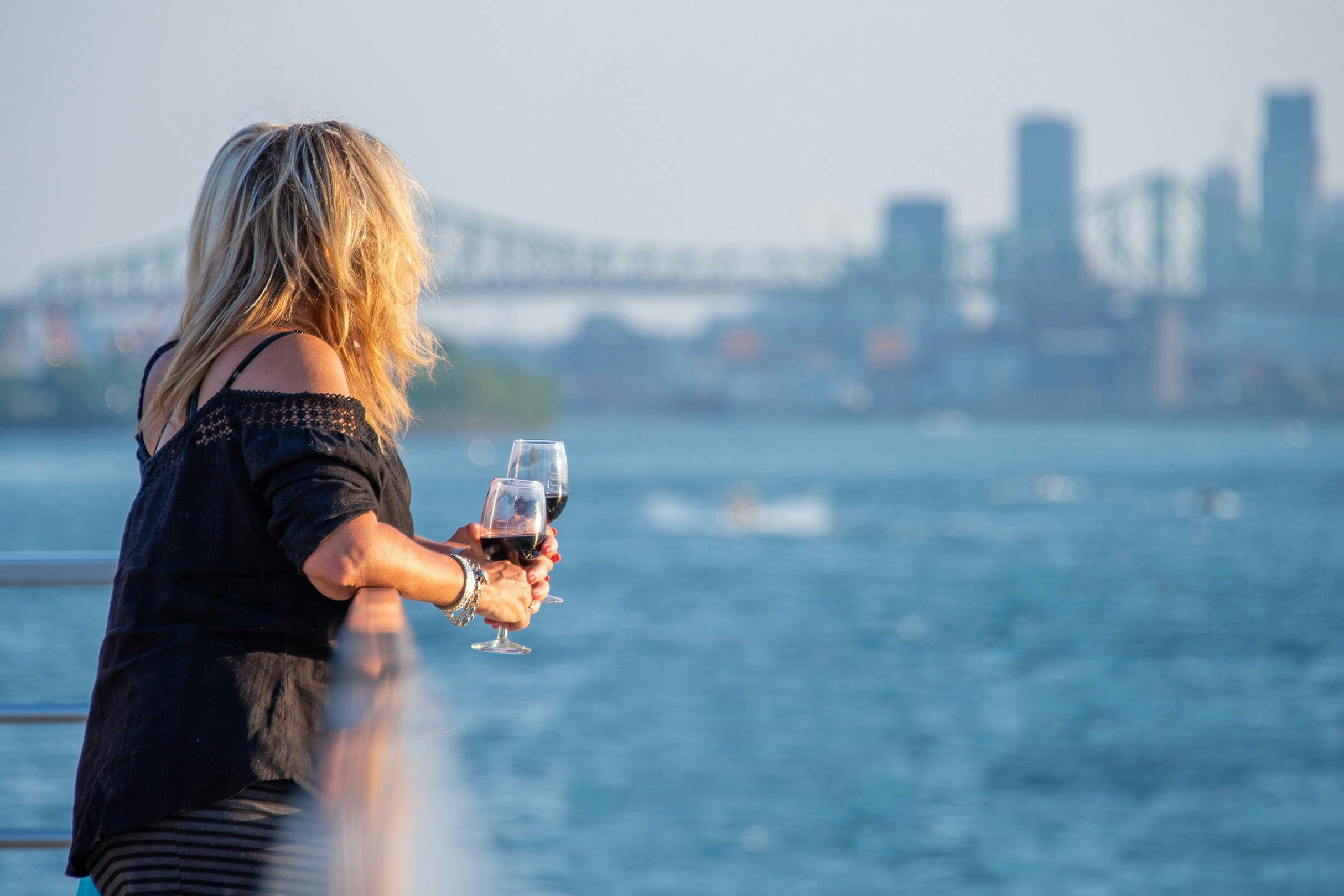 A woman holding two wine glasses stands by the water with a bridge and cityscape in the background.