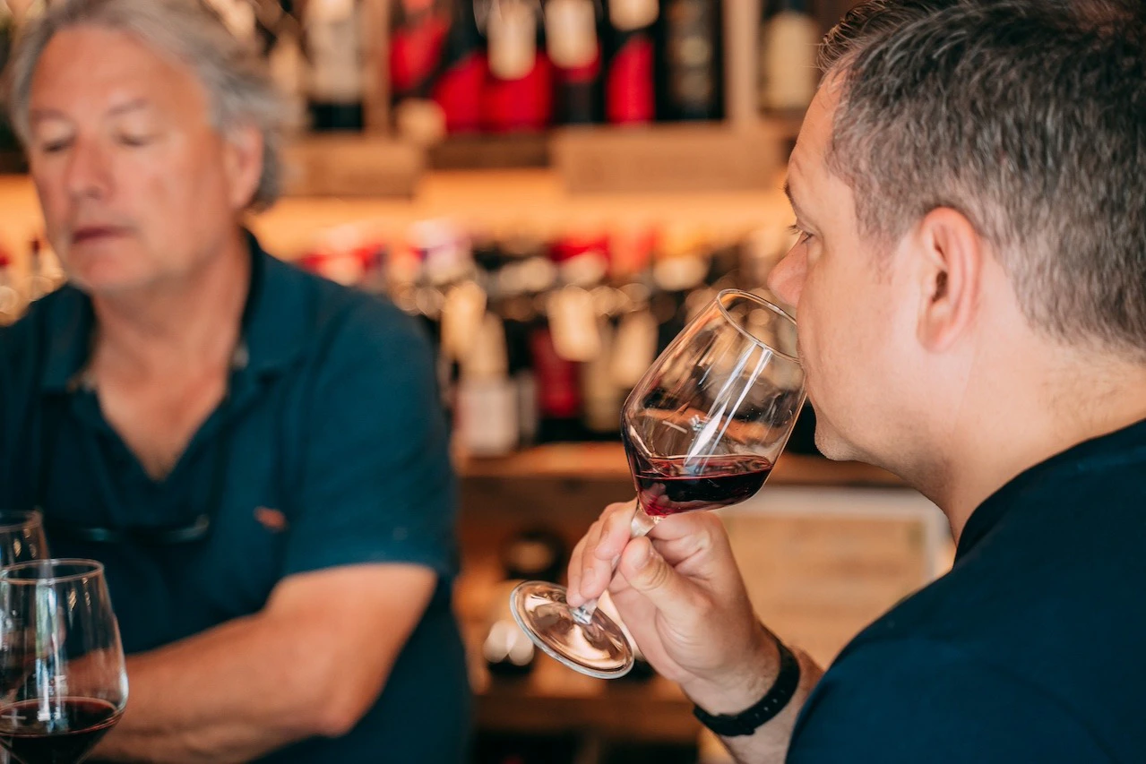Two people in a wine shop, one holding a glass of red wine near their face, shelves of wine bottles in the background.