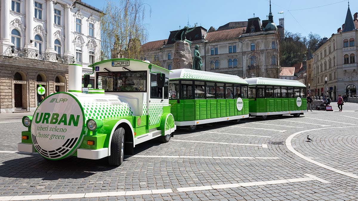 Zeleno-bílý turistický vláček zaparkovaný na dlážděném náměstí před historickými budovami.
