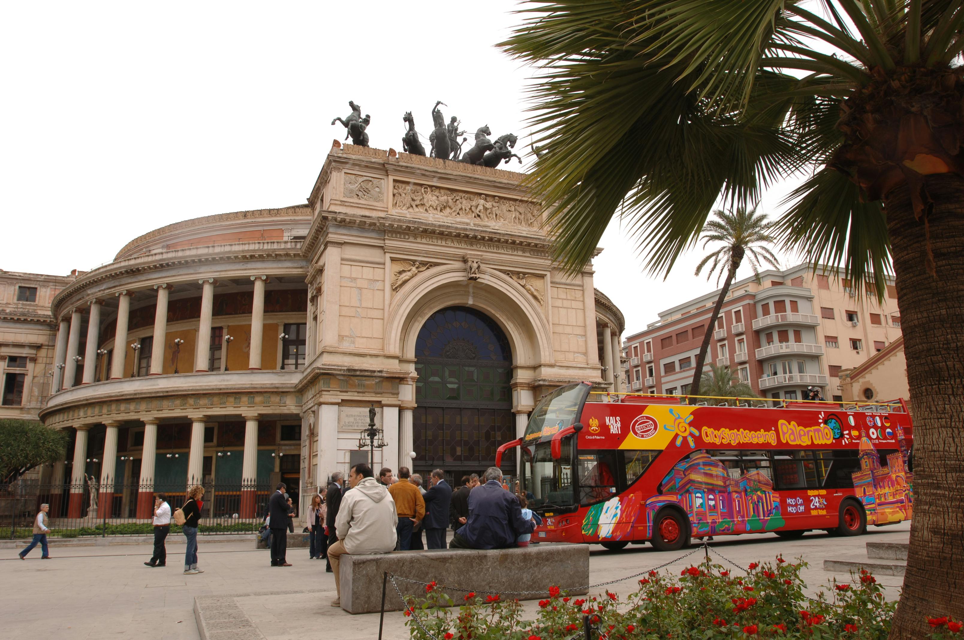 People gathered near an ornate building with statues on top. A bright red double-decker tour bus is parked in the foreground.