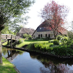 En stuga med halmtak står bland gröna träd och buskar vid en smal kanal, med en träbro som korsar vattnet.