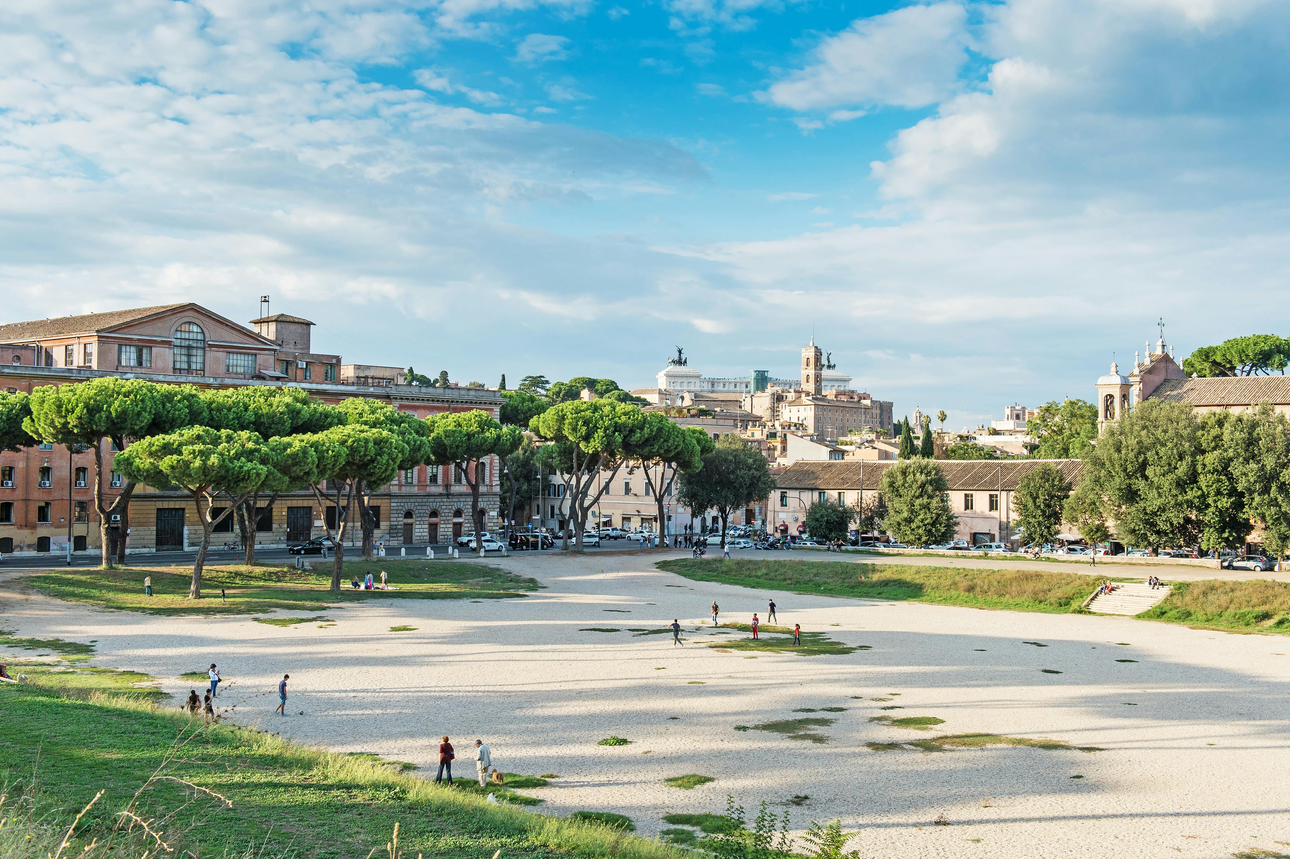 Diverse persone passeggiano e si rilassano in una distesa aperta e rocciosa circondata da alberi ed edifici storici sotto un cielo azzurro e parzialmente nuvoloso.
