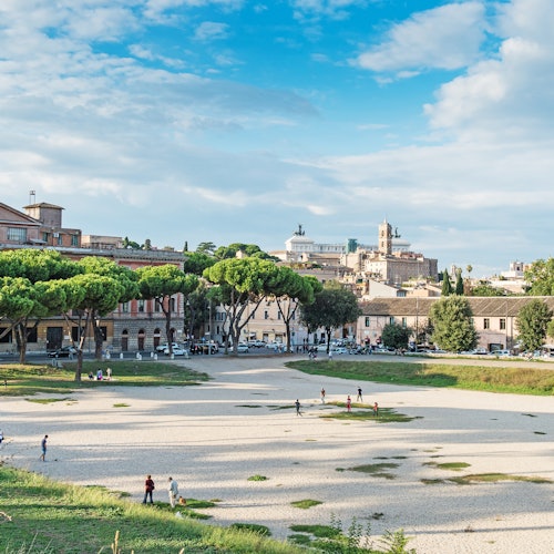 Diverse persone passeggiano e si rilassano in una distesa aperta e rocciosa circondata da alberi ed edifici storici sotto un cielo azzurro e parzialmente nuvoloso.