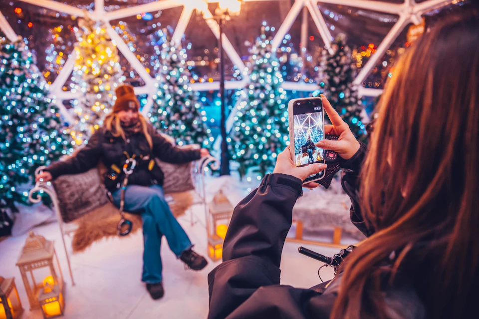 A person takes a photo of another person seated in a festive setting with decorated trees and bright lights.