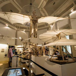 Människor observerar flygande maskinmodeller och diagram i ett väl upplyst museumsutställningsrum med högt i tak och invecklad arkitektur.