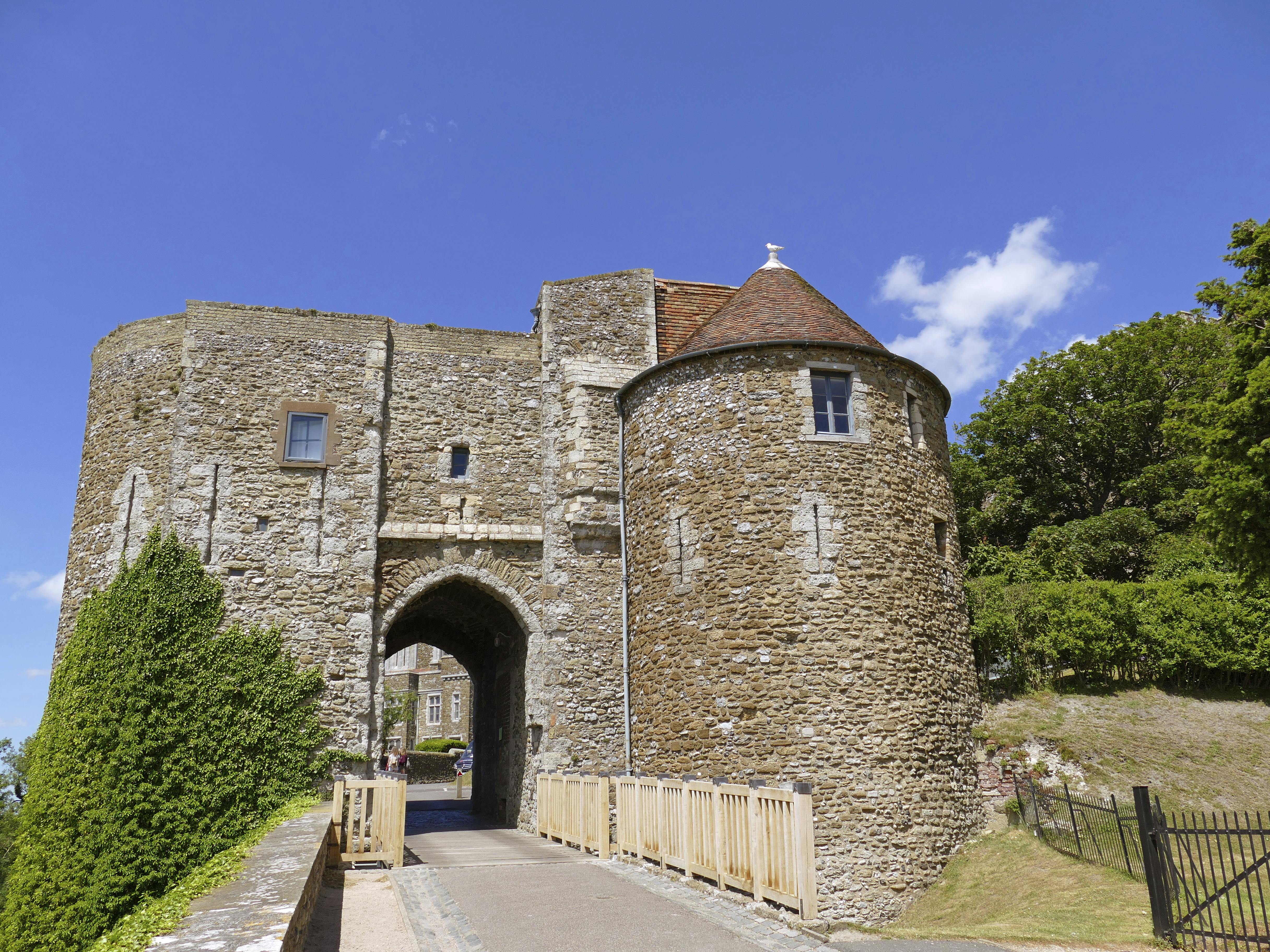 Μια πέτρινη είσοδος κάστρου με στρογγυλό πυργίσκο, ξύλινη γέφυρα και γύρω πράσινο κάτω από έναν καταγάλανο ουρανό.