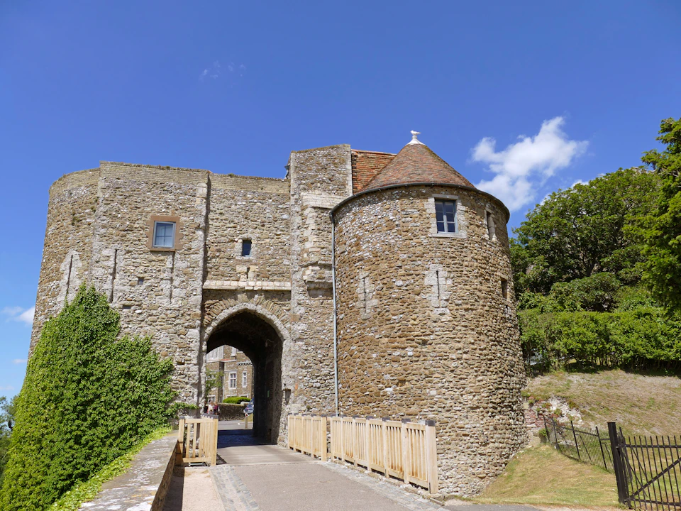 A stone castle entrance with a round turret, a wooden bridge, and surrounding greenery under a clear blue sky.