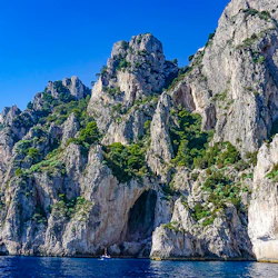 Branta, steniga klippor täckta av grönska reser sig över ett lugnt blått hav under en klar himmel. En liten båt ligger nära basen av klipporna.