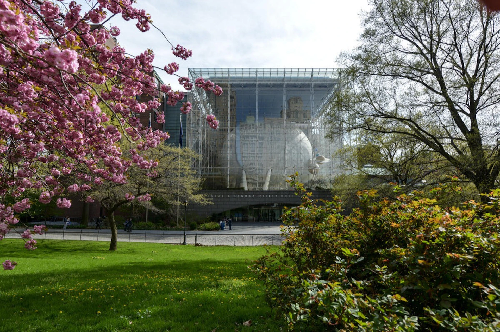 A glass building with a large sphere inside, surrounded by greenery and blooming trees on a sunny day.