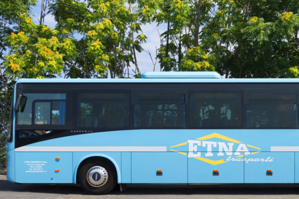 A blue tour bus labeled "ETNA" parked in front of trees with yellow flowers.