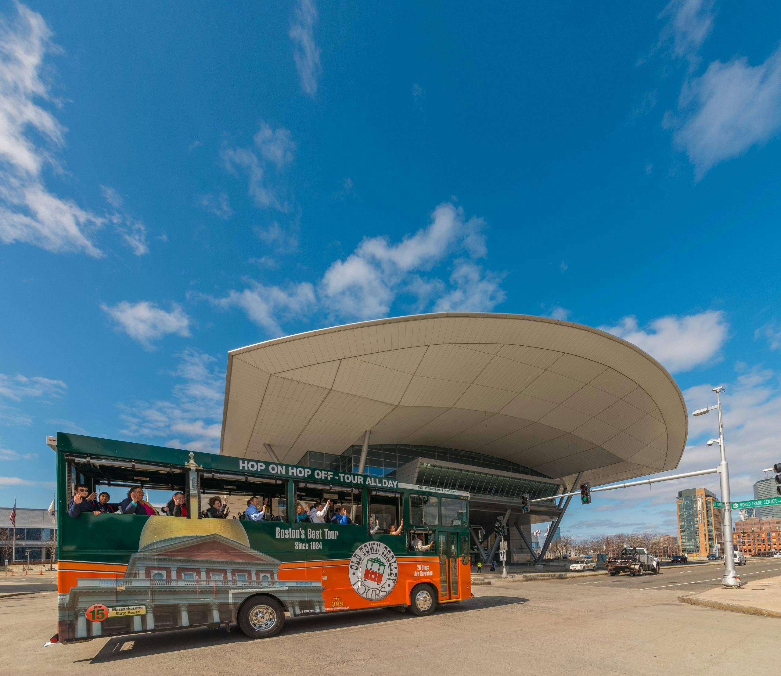 Een groene en oranje tourbus met passagiers passeert een groot, modern gebouw onder een blauwe lucht met wolken.
