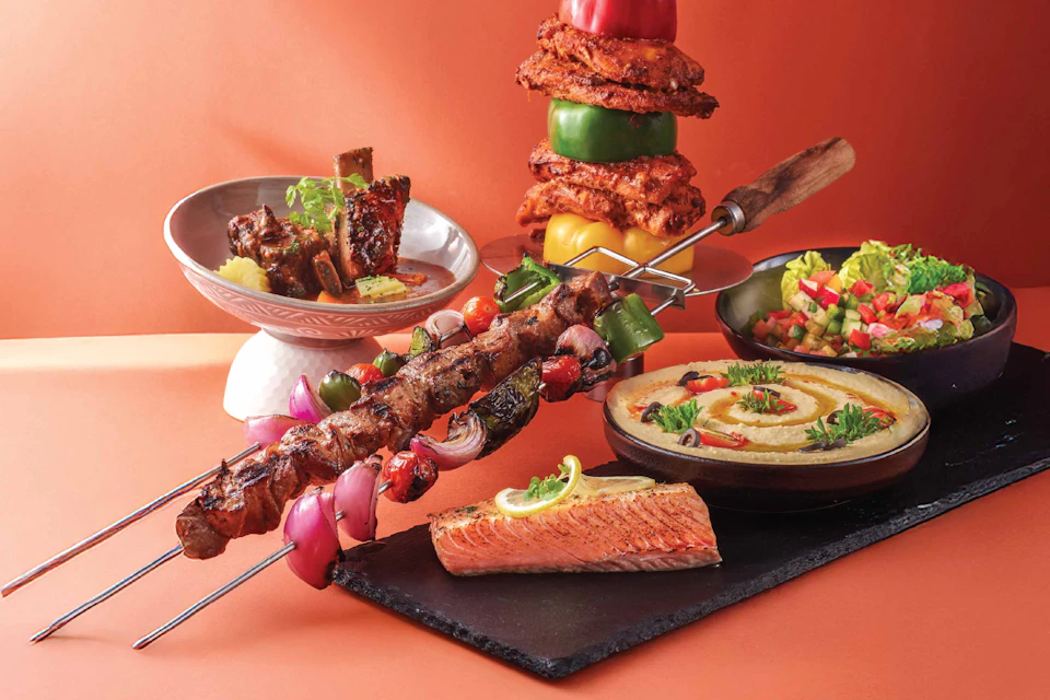 A variety of grilled and fresh dishes, including kebabs, hummus, grilled salmon, and vegetables, arranged on a table.