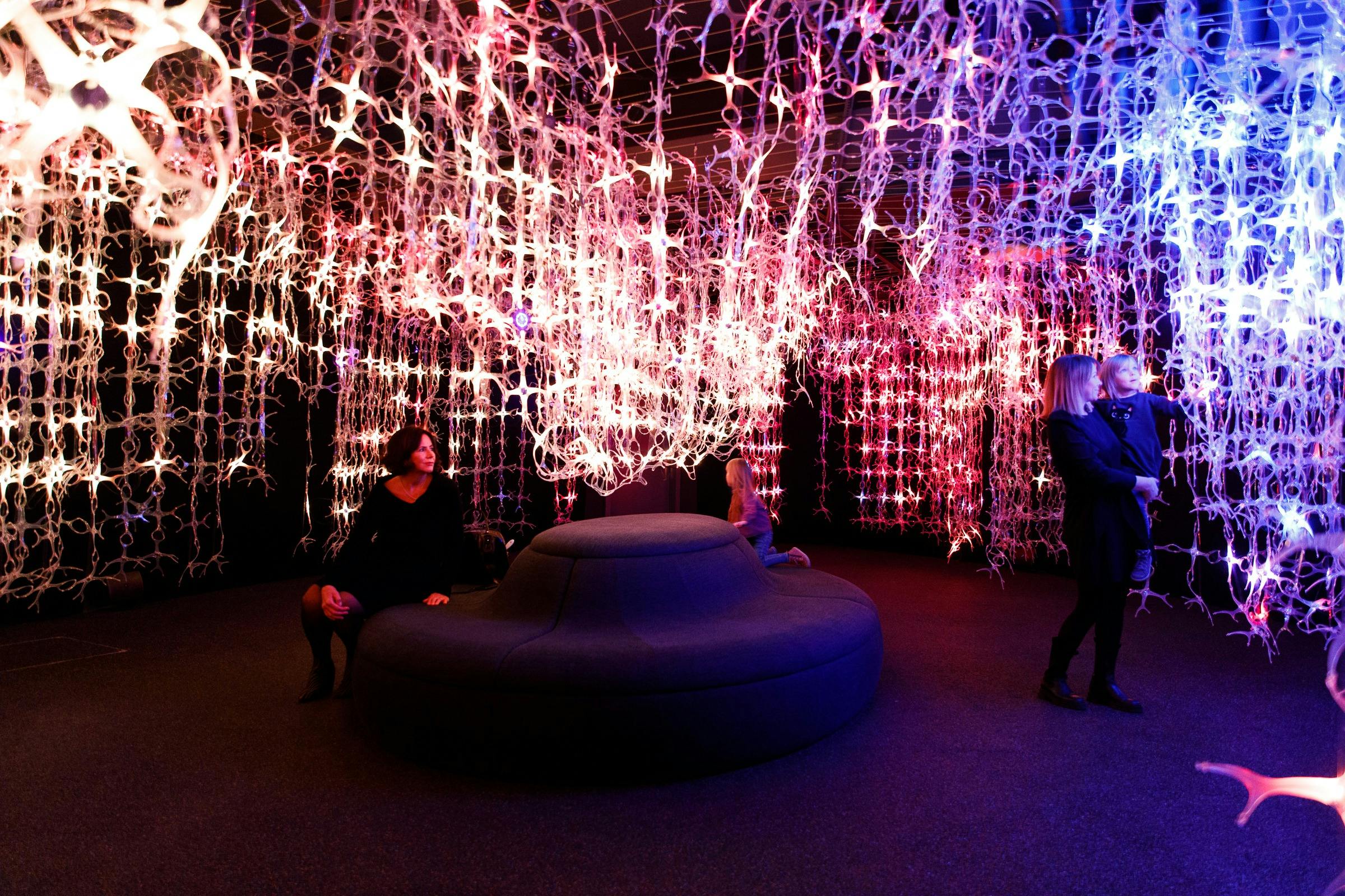 Άνθρωποι κάθονται και στέκονται γύρω από ένα σκοτεινό δωμάτιο με φωτιζόμενες, περίπλοκες, κρεμαστές εγκαταστάσεις σε διάφορα χρώματα.