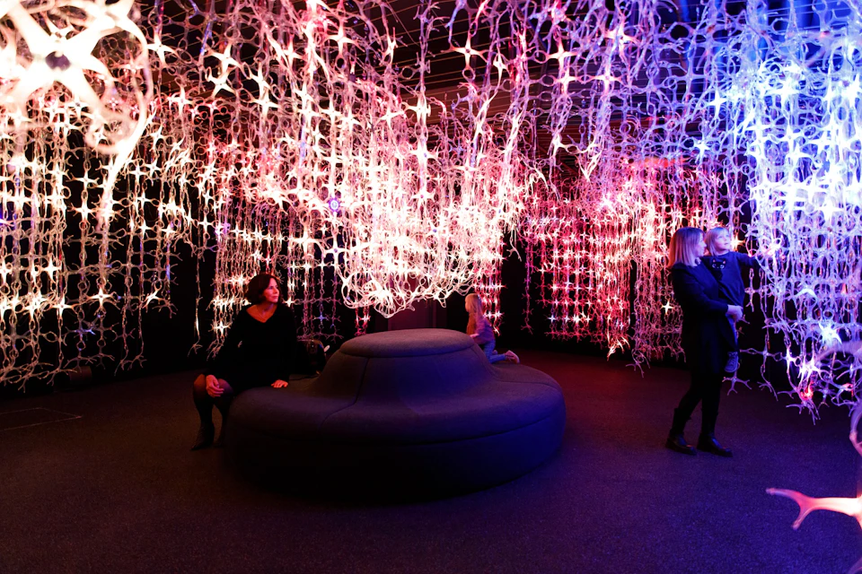 People sitting and standing around a dark room with illuminated, intricate, hanging installations in various colors.