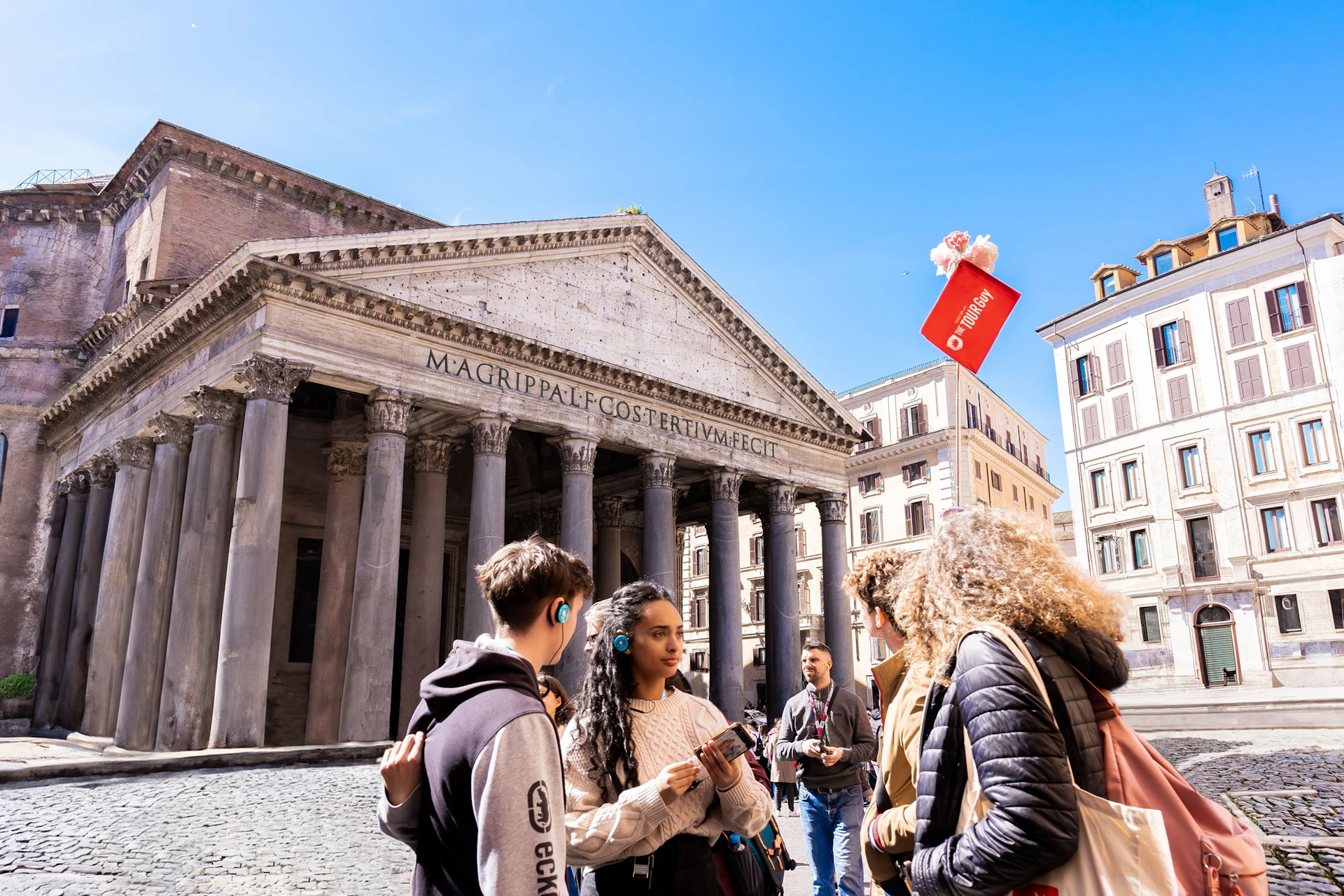 A group of tourists is standing in front of the Pantheon in Rome on a sunny day. A red city map is floating above them.