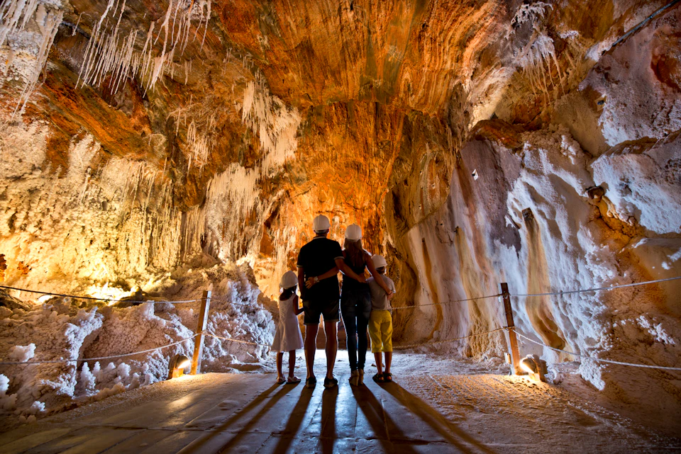A family wearing helmets stands inside a brightly lit cave, admiring the stalactites and stalagmites.