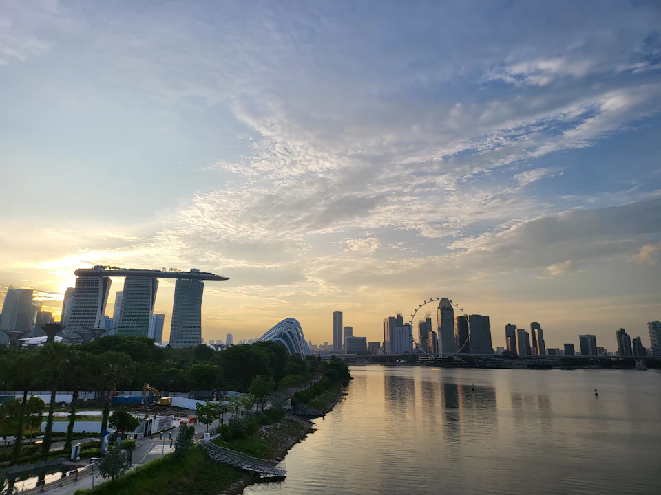 A city skyline at sunset with a waterfront, iconic buildings, and a Ferris wheel under a partly cloudy sky.