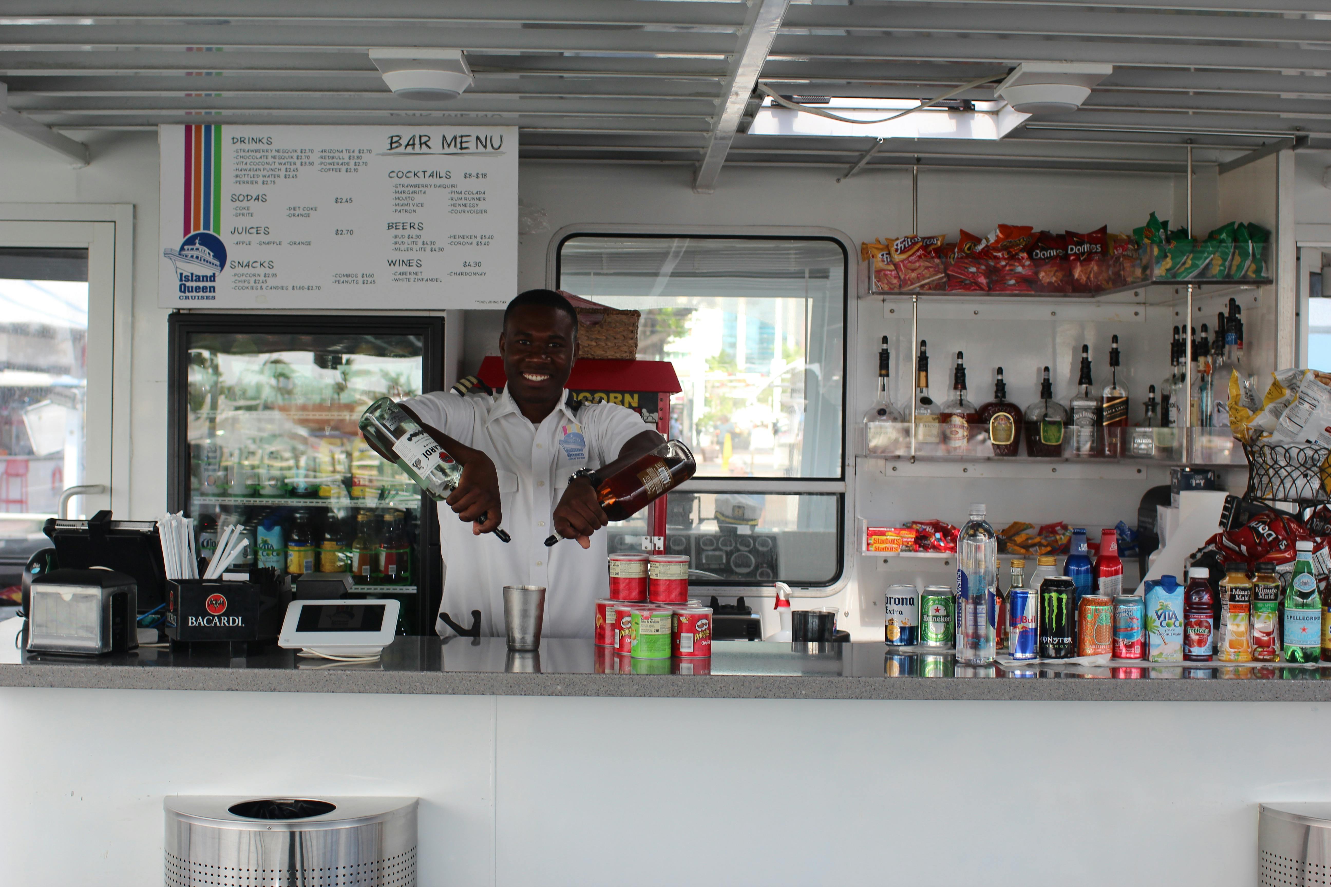 Χαμογελαστός μπάρμαν που αναμιγνύει ποτά πίσω από ένα μπαρ με διάφορα ποτά, σνακ και ένα μενού στο παρασκήνιο.