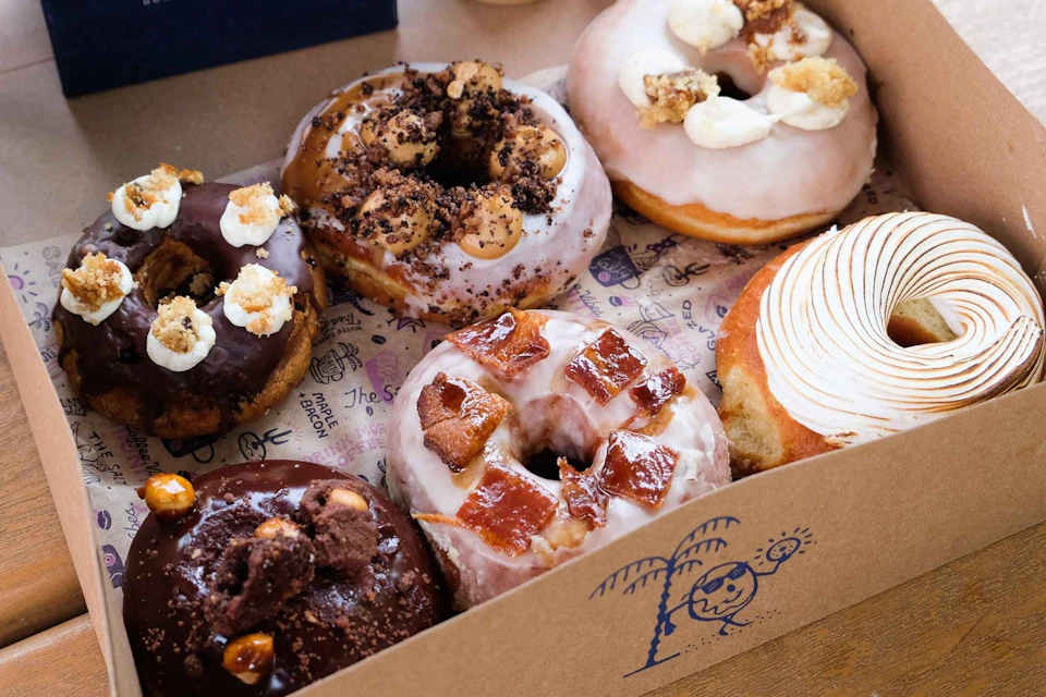 A box containing six assorted gourmet doughnuts, each topped with different glazes, crumbs, and toppings.